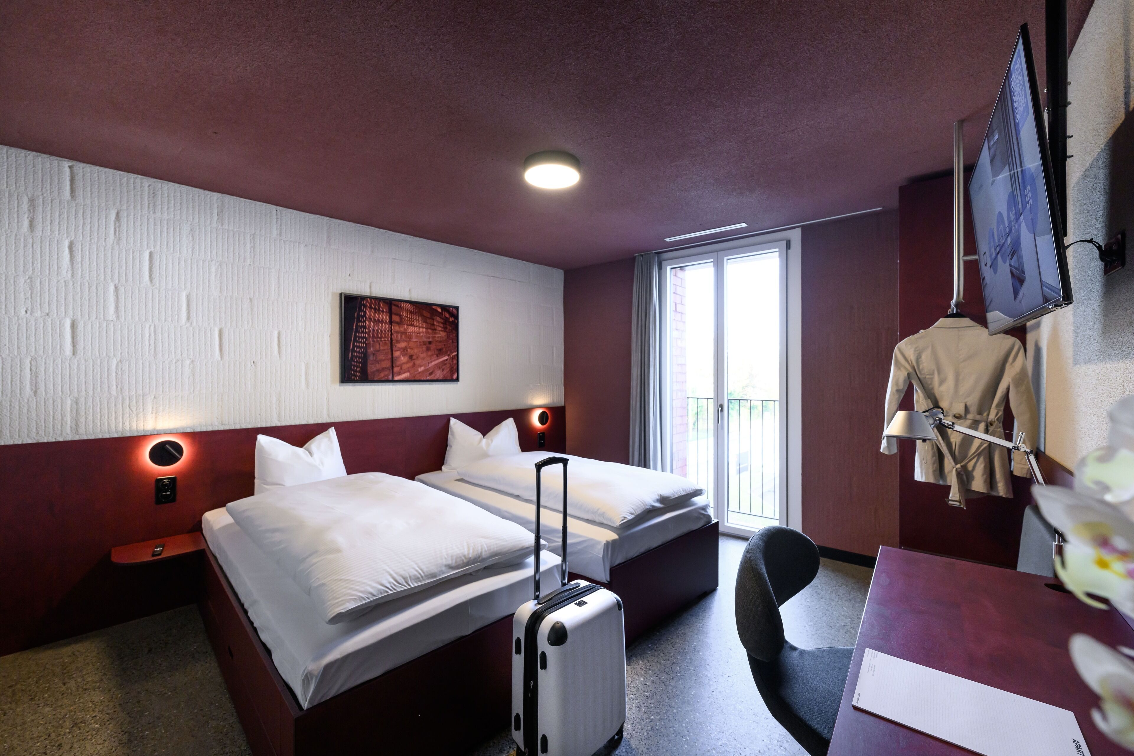 Habitación de hotel con dos camas, un escritorio, una silla, una maleta, un perchero y un televisor.