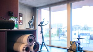 Une salle de sport équipée de divers appareils d’exercice, d’un tapis de course et d’un vélo d’appartement. On trouve des bouteilles d’eau et des tapis enroulés sur une étagère.