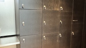 Une rangée de casiers avec des poignées et des serrures métalliques, une petite étagère sur le côté gauche, et un sol texturé composé de carreaux orange et beige.