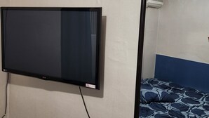 Ein Zimmer mit flachbildfernseher an der Wand, einem Bett mit gemustertem Überwurf und einem Spiegel, der die Deckenleuchte des Zimmers reflektiert.