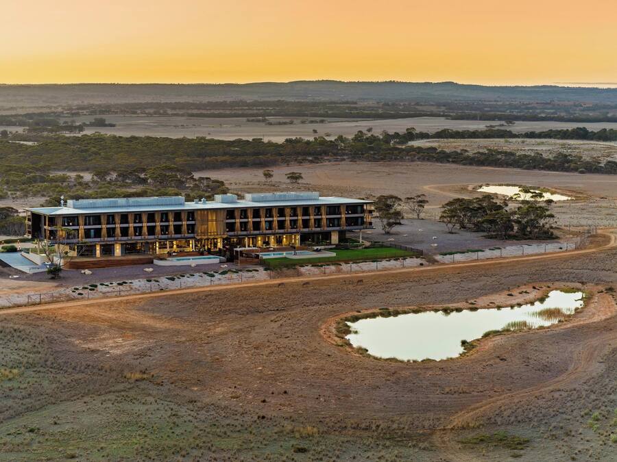Un edificio moderno con techo plano, rodeado por un paisaje árido con poca vegetación y algunos cuerpos de agua.