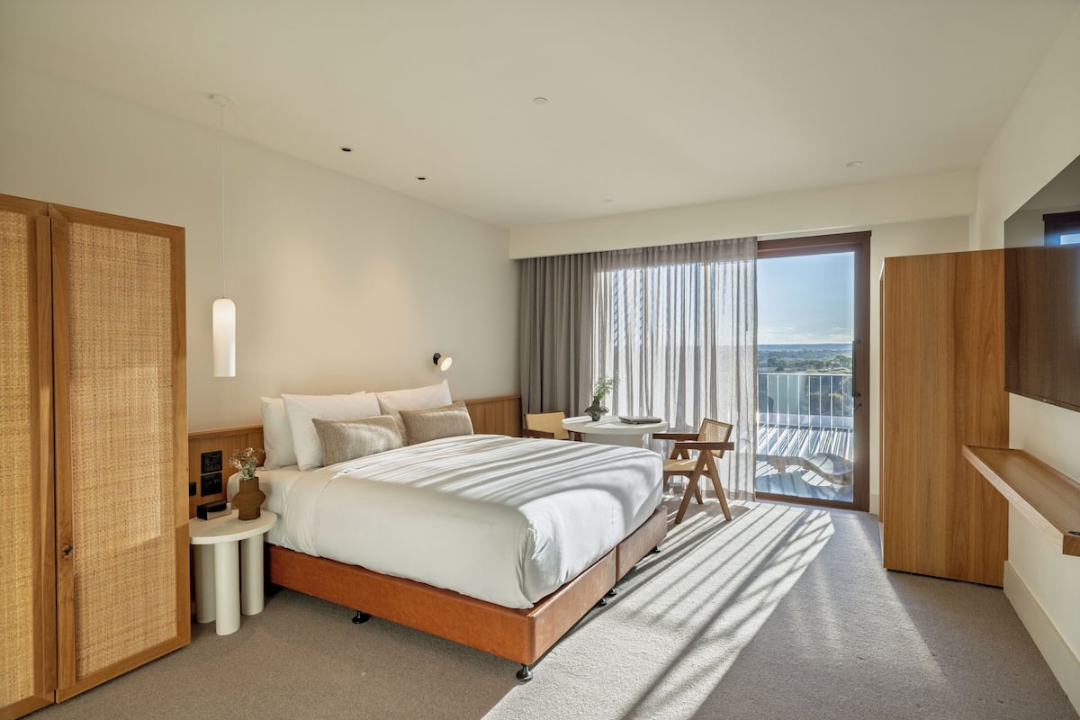 A modern hotel room with a large bed, a small round table, a TV, and a balcony with a view.