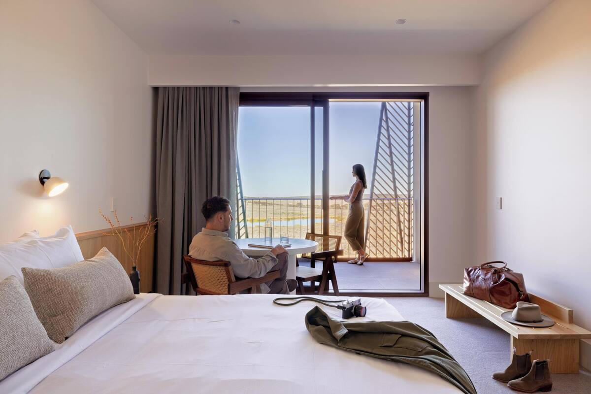 A hotel room with a bed, a table, a chair, a bench, a suitcase, a camera, and a view of the outside.