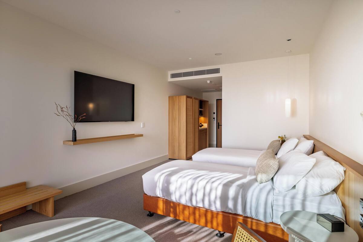 A modern hotel room with a large bed, a TV mounted on the wall, a wooden bench, and a small table.