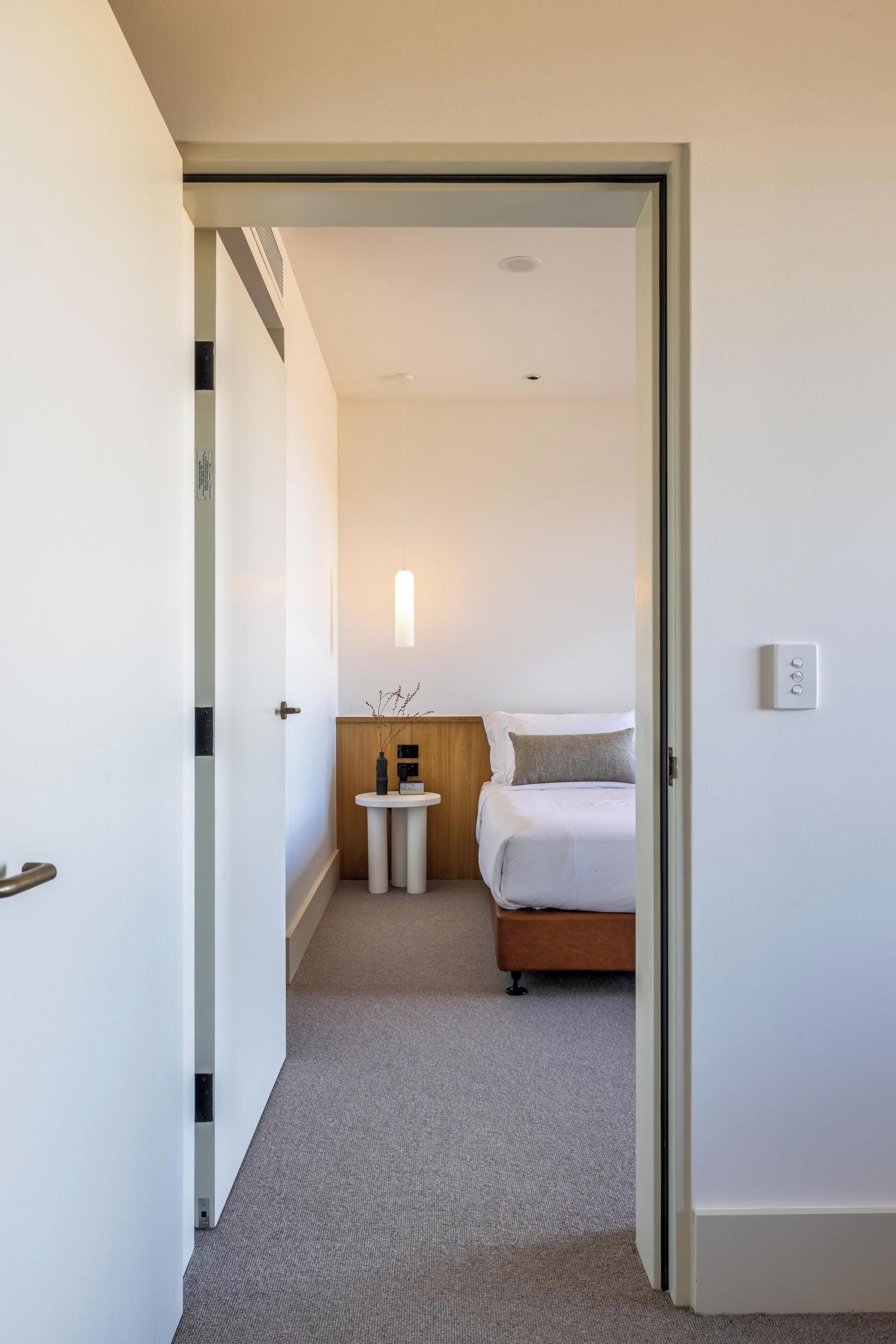 Hotellihuone, jossa on sänky, yöpöytä ja pieni seinään kiinnitetty valaisin.