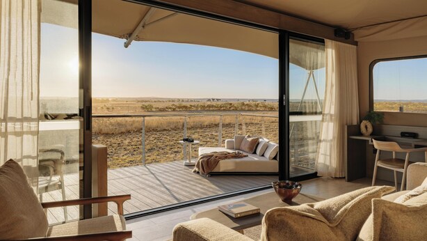 A modern interior with a large glass door leading to an outdoor deck, a sofa, a dining area, and a view of the landscape. - Monarto Safari Resort (Monarto)