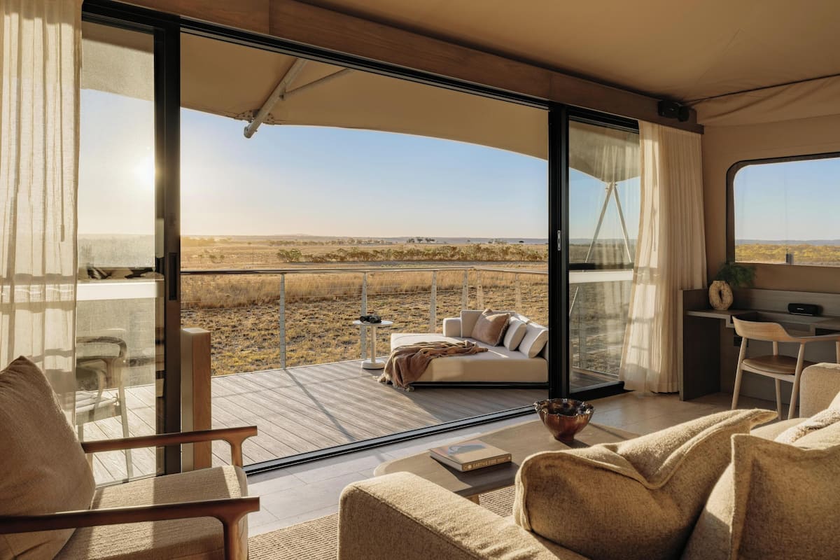 A modern interior with a large glass door leading to an outdoor deck, a sofa, a dining area, and a view of the landscape.