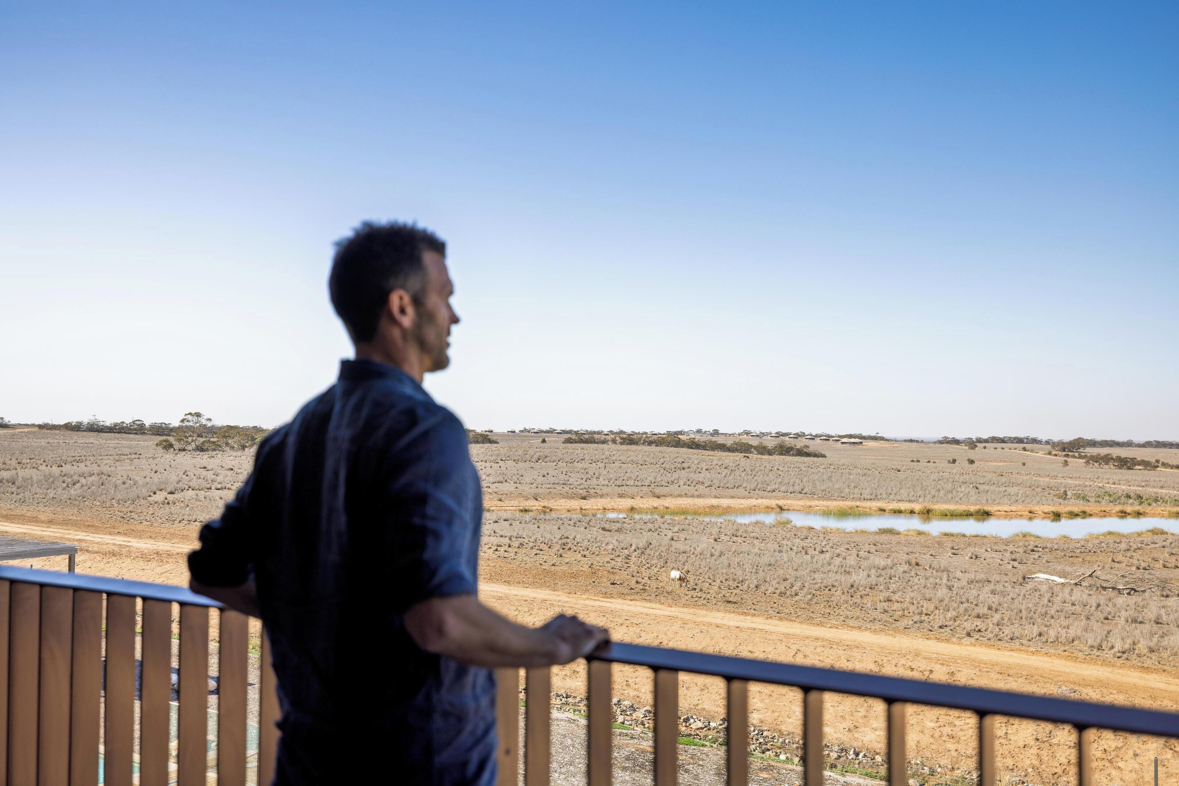 A man standing on a balcony overlooking a dry landscape with sparse vegetation and a dirt road.