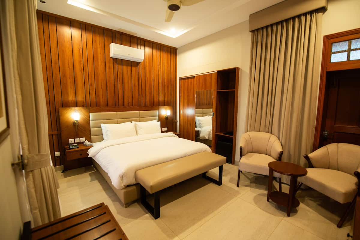 Une chambre d’hôtel avec un grand lit, une tête de lit en bois, un banc, deux fauteuils, une petite table, un miroir et une armoire.