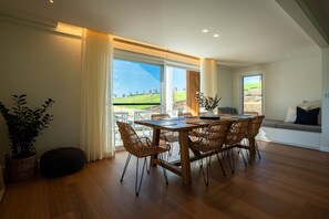 Un área de comedor con mesa de madera y sillas de mimbre, ventanas grandes con cortinas y vistas a un paisaje verde.