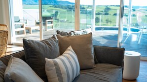 Ein modernes Wohnzimmer mit einer Couch, Kissen und einem weißen, zylindrischen Objekt, vermutlich ein Lautsprecher, mit einer großen Glasschiebetür zu einem Außenbereich mit Blick auf eine grüne Landschaft.