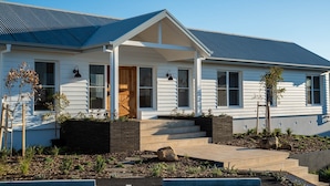 A modern two-story house with a covered porch, a brick retaining wall, and a landscaped garden with plants and a rock feature. - Luxury Coastal Vineyard Escape | Fig View | Crooked River Estate (Gerringong)