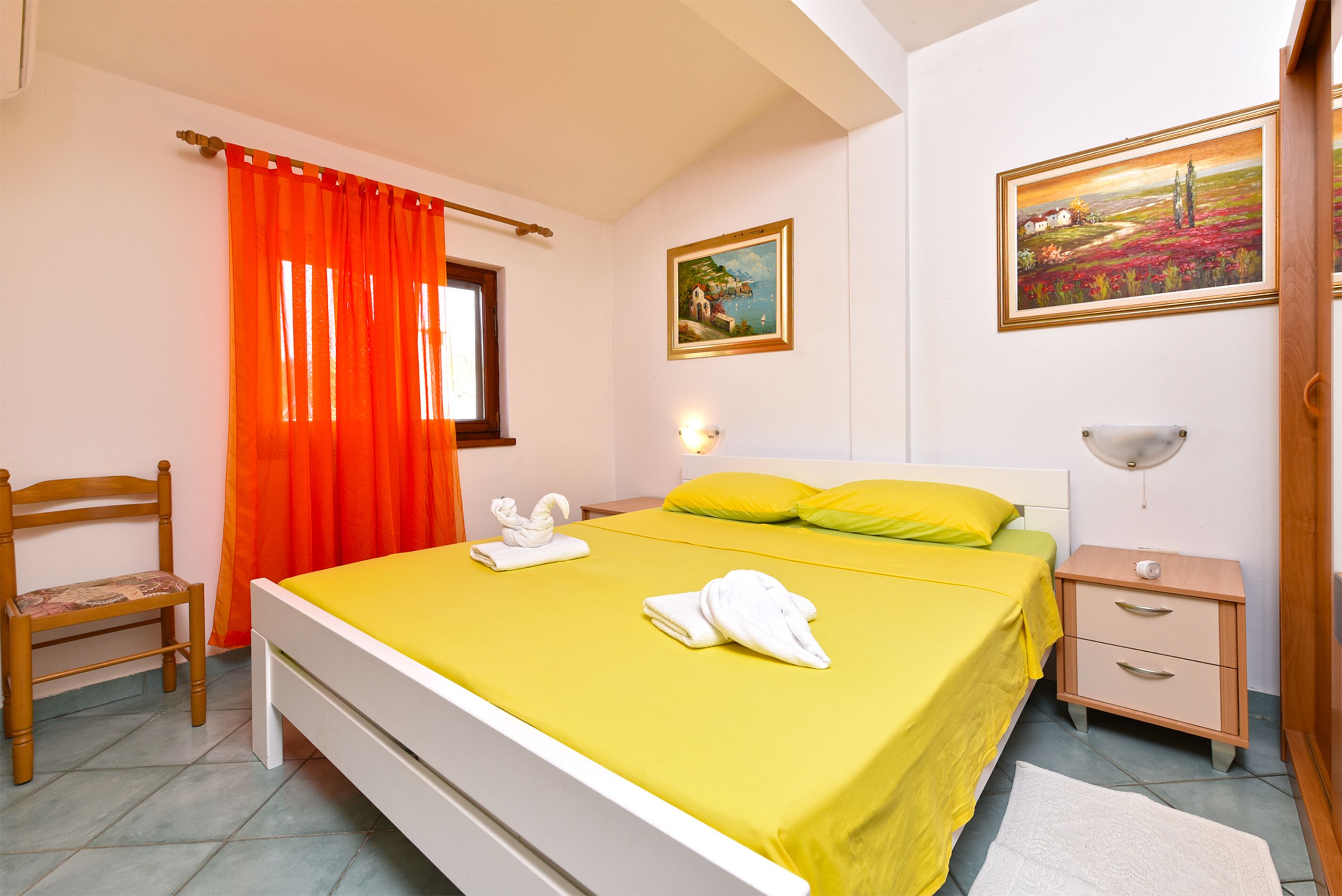 A bedroom with a bed, a chair, a nightstand, and two paintings on the wall.