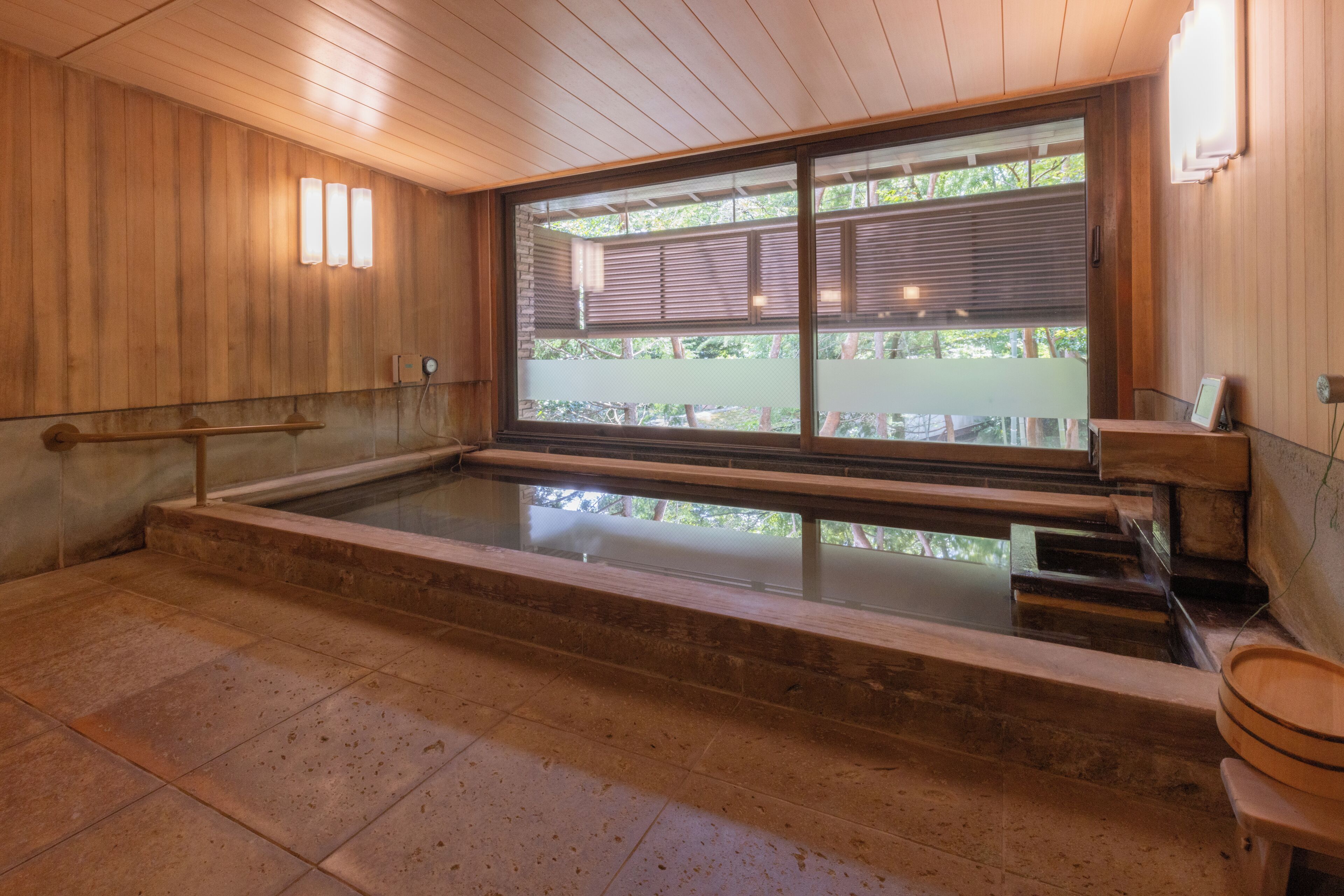 A traditional Japanese onsen with a wooden interior, a stone hot spring bath, and a view through a shoji screen.