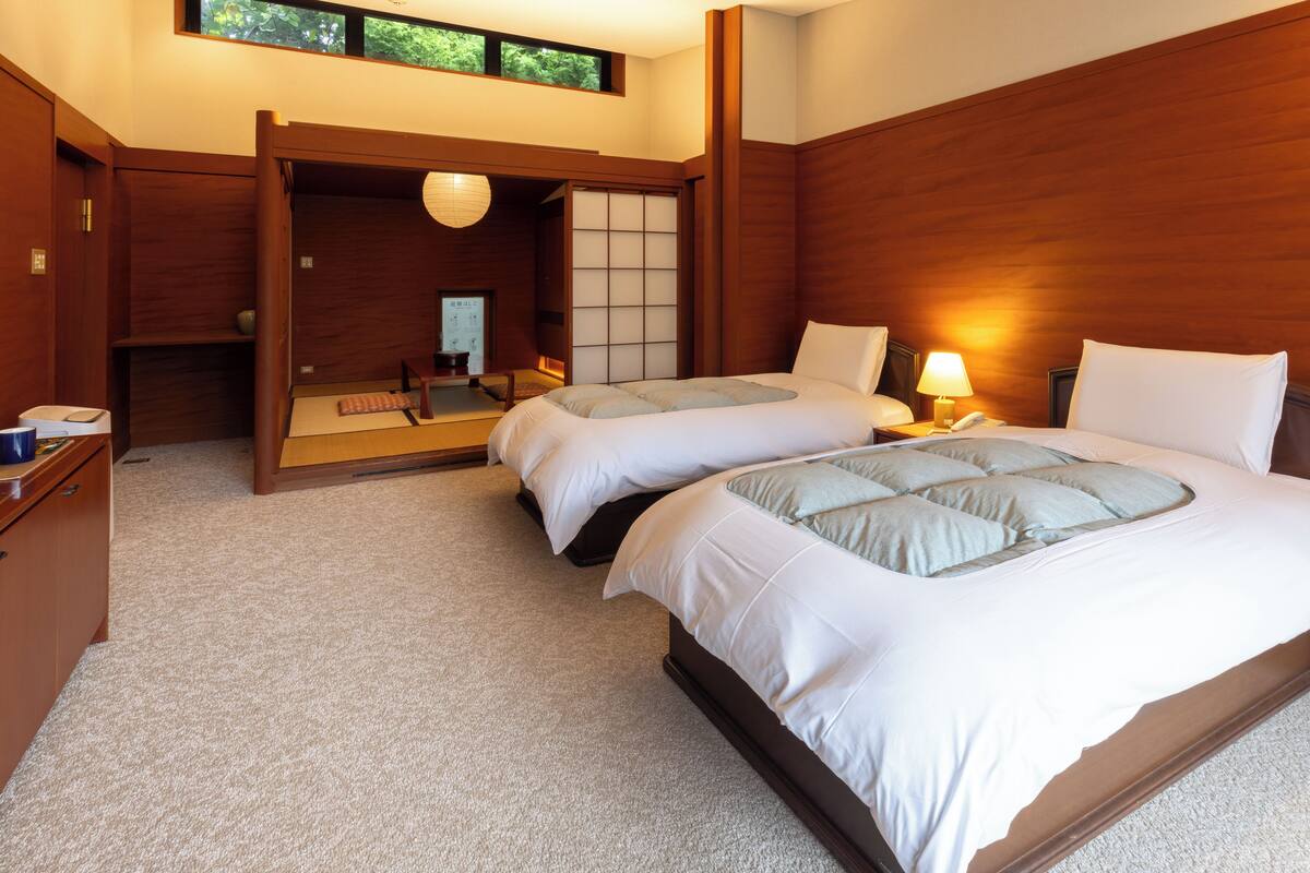 Quarto de hotel com duas camas, cabeceiras de madeira, uma porta deslizante tradicional japonesa e uma pequena escrivaninha com abajur.