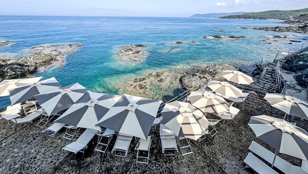 Een strand met helderblauw water, rotsachtig terrein en ligbedden en parasols.