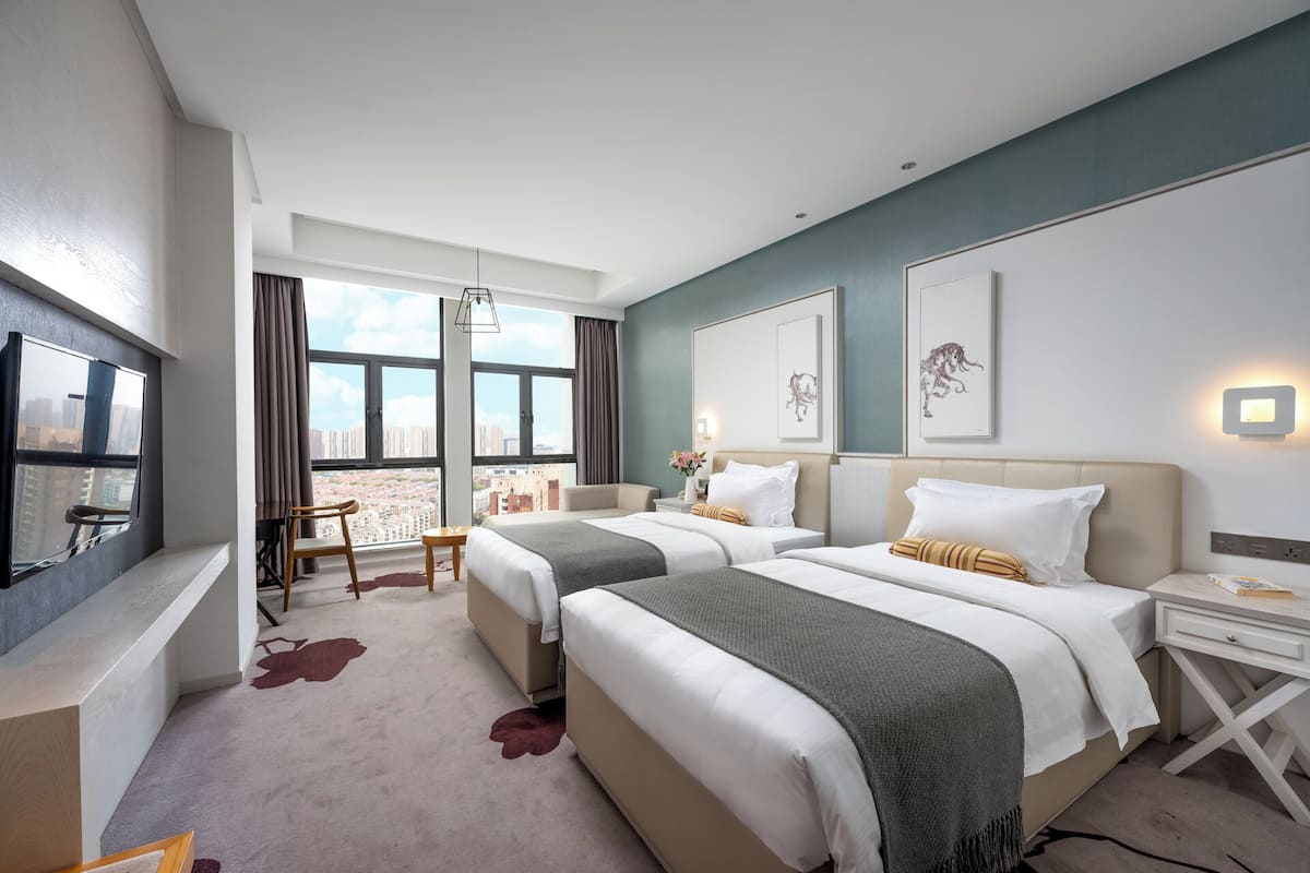 A hotel room with two beds, a desk, a chair, a TV, and large windows offering a city view.