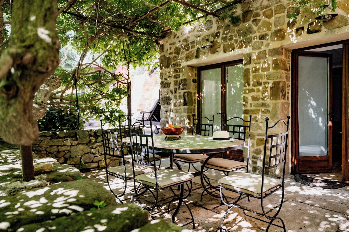 A stone building with a round table and chairs, a stone wall, and a tree in the foreground.