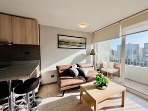 A modern living space with a sofa, coffee table, and a view of a cityscape through large windows. - 1BR La Florida - Free Parking, Pool & Metro (La Florida)