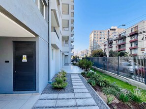 A residential building with a gated entrance, a narrow pathway lined with potted plants, and a fenced area with parked cars. - 1BR La Florida - Free Parking, Pool & Metro (La Florida)