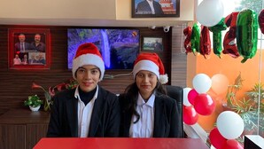 Deux personnes portant des bonnets de Père Noël sont assises à une table recouverte d’une nappe rouge. Un téléviseur diffuse une image de cascade, et un cadre représentant un juge est accroché au mur.