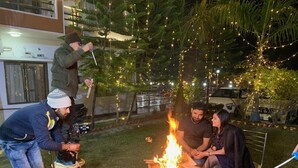 Un groupe de personnes est réuni autour d’un feu de camp dans un cadre extérieur, avec des guirlandes lumineuses.