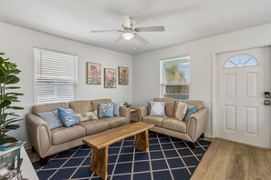 A living room with two sofas, a coffee table, and a ceiling fan. There are three framed pictures on the wall, and a door with a window above it. - Updated 3-bedroom, 2 bath coastal cottage across from the beach UNIT A (Fernandina Beach)