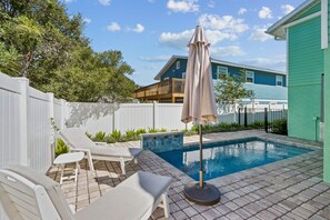 A swimming pool with lounge chairs and an umbrella, surrounded by a white fence and greenery.