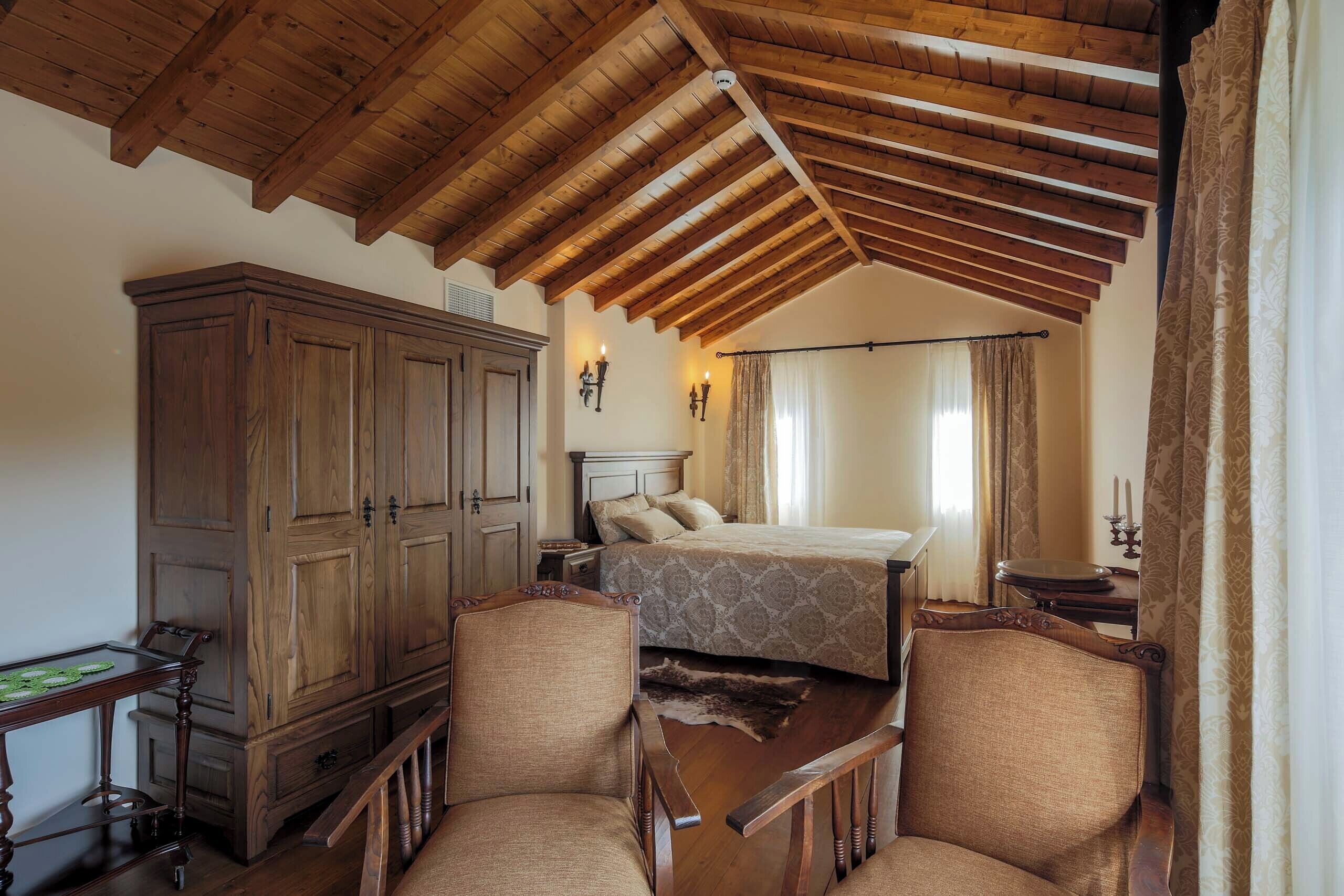 A bedroom with a wooden ceiling, a large wardrobe, two upholstered chairs, a bed with patterned bedding, and a nightstand with a lamp.