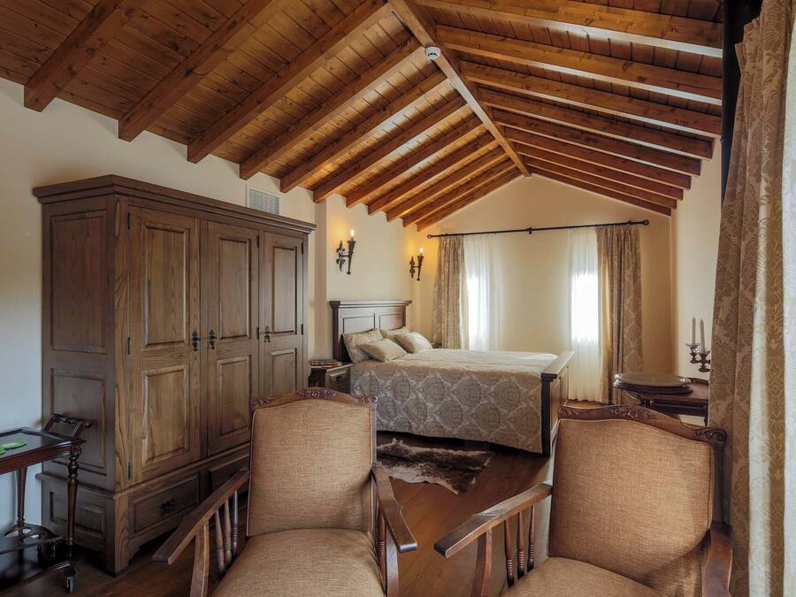 A bedroom with a wooden ceiling, a large wardrobe, two upholstered chairs, a bed with patterned bedding, and a nightstand with a lamp.