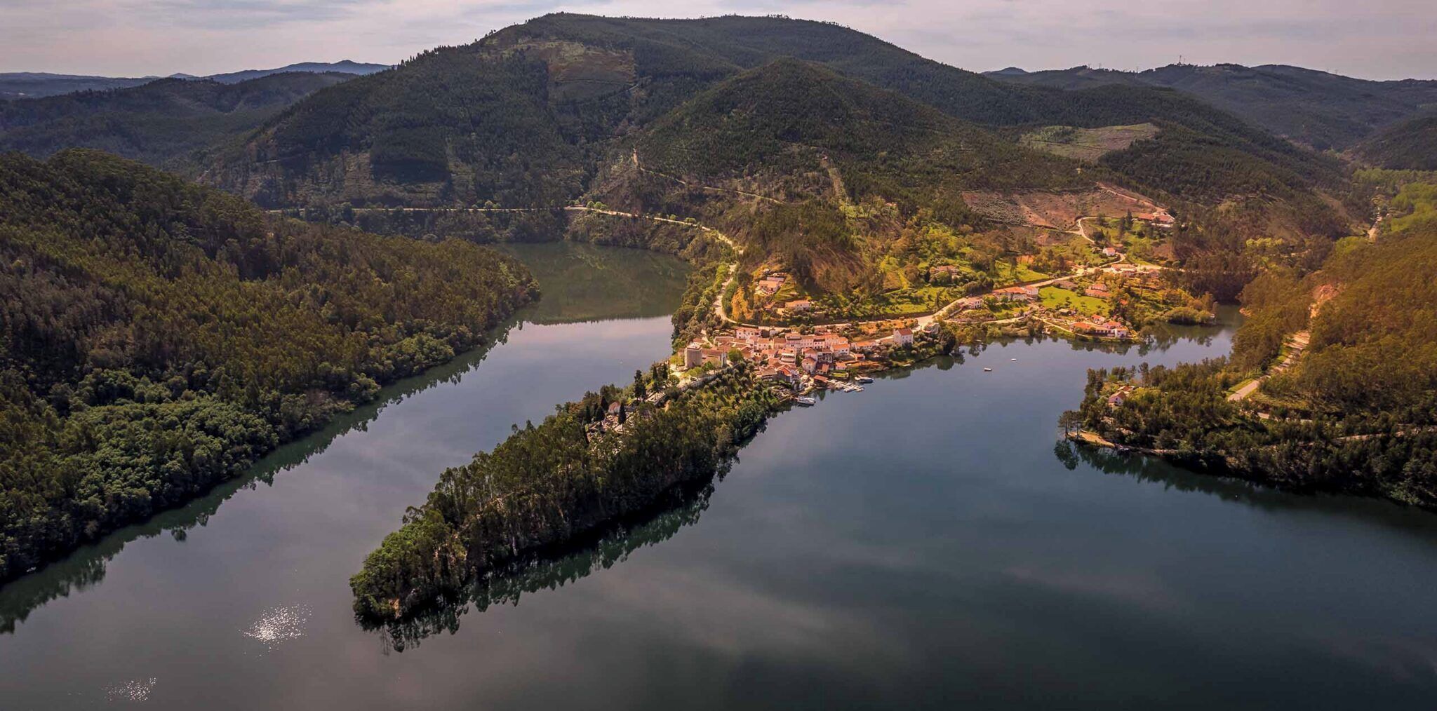 Vista panoràmica d'una vila a la riba d'un riu, amb una església destacada i turons boscosos al voltant.