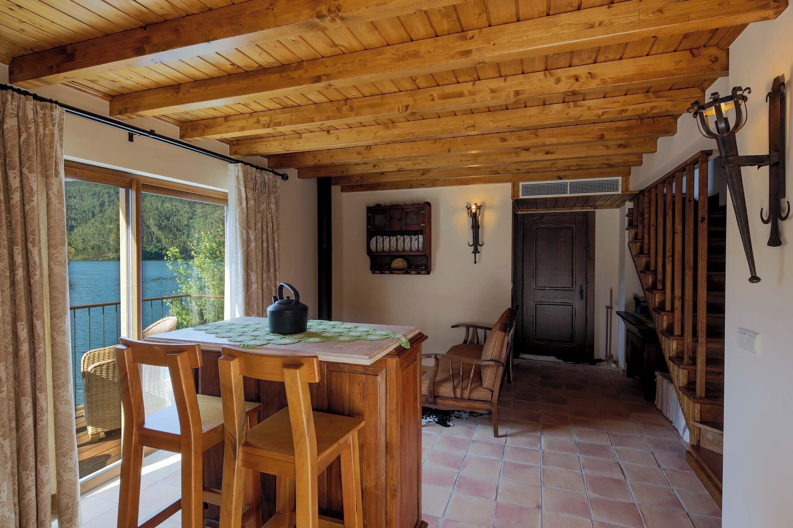 A wooden dining table with chairs, a view of a lake through a large window, a wooden staircase, and a door with a dark frame.
