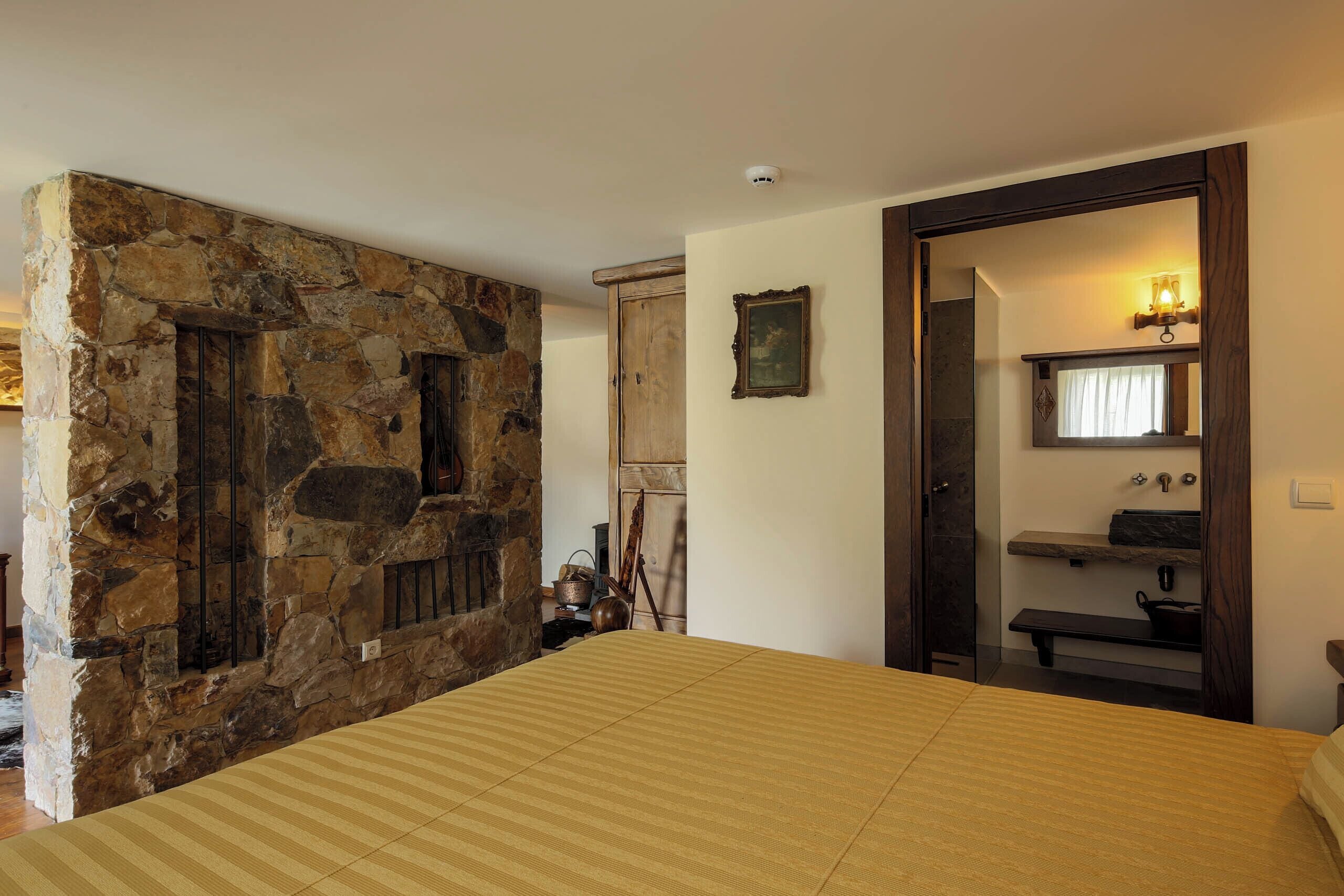 A bedroom with a stone wall, a bed with a striped bedspread, a wooden door, a mirror, and a shelf with toiletries.