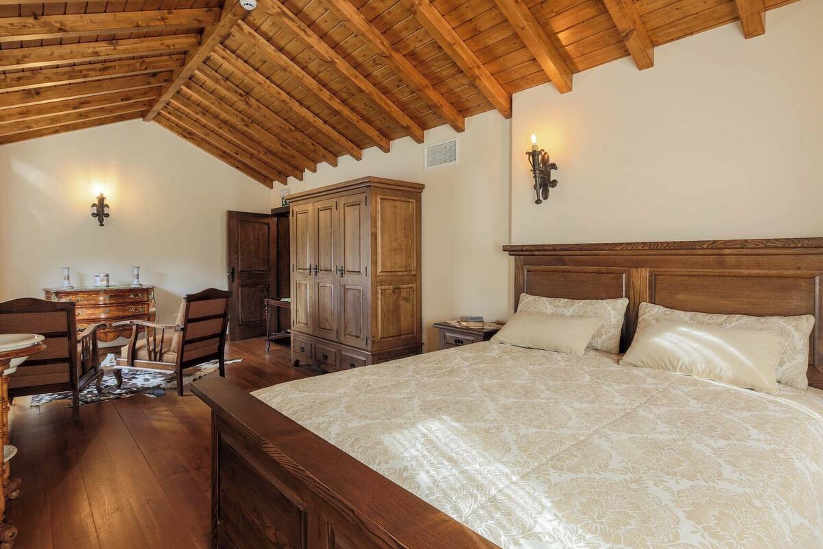 Une chambre avec un plafond en bois, un grand lit recouvert d’une couvre-lit à motifs, une commode en bois et deux lampes fixées au mur.