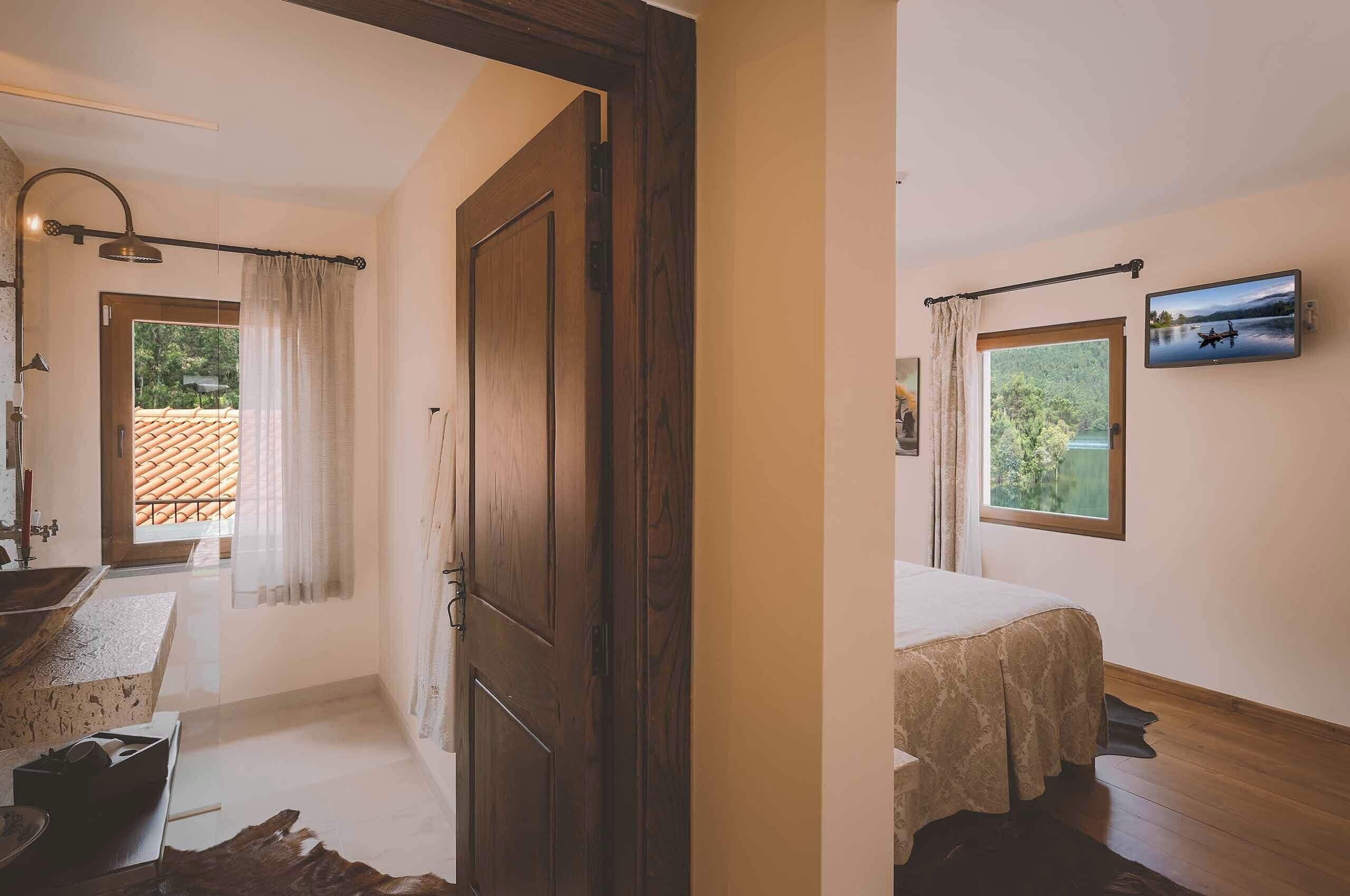 Une pièce avec une porte en bois, un lit recouvert d’une couverture beige, une fenêtre donnant sur la verdure et un téléviseur fixé au mur.