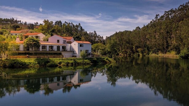 Une propriété située au bord d’un lac, avec un bâtiment blanc doté de nombreux balcons et d’un toit rouge, entourée d’une forêt dense et d’un lac paisible.