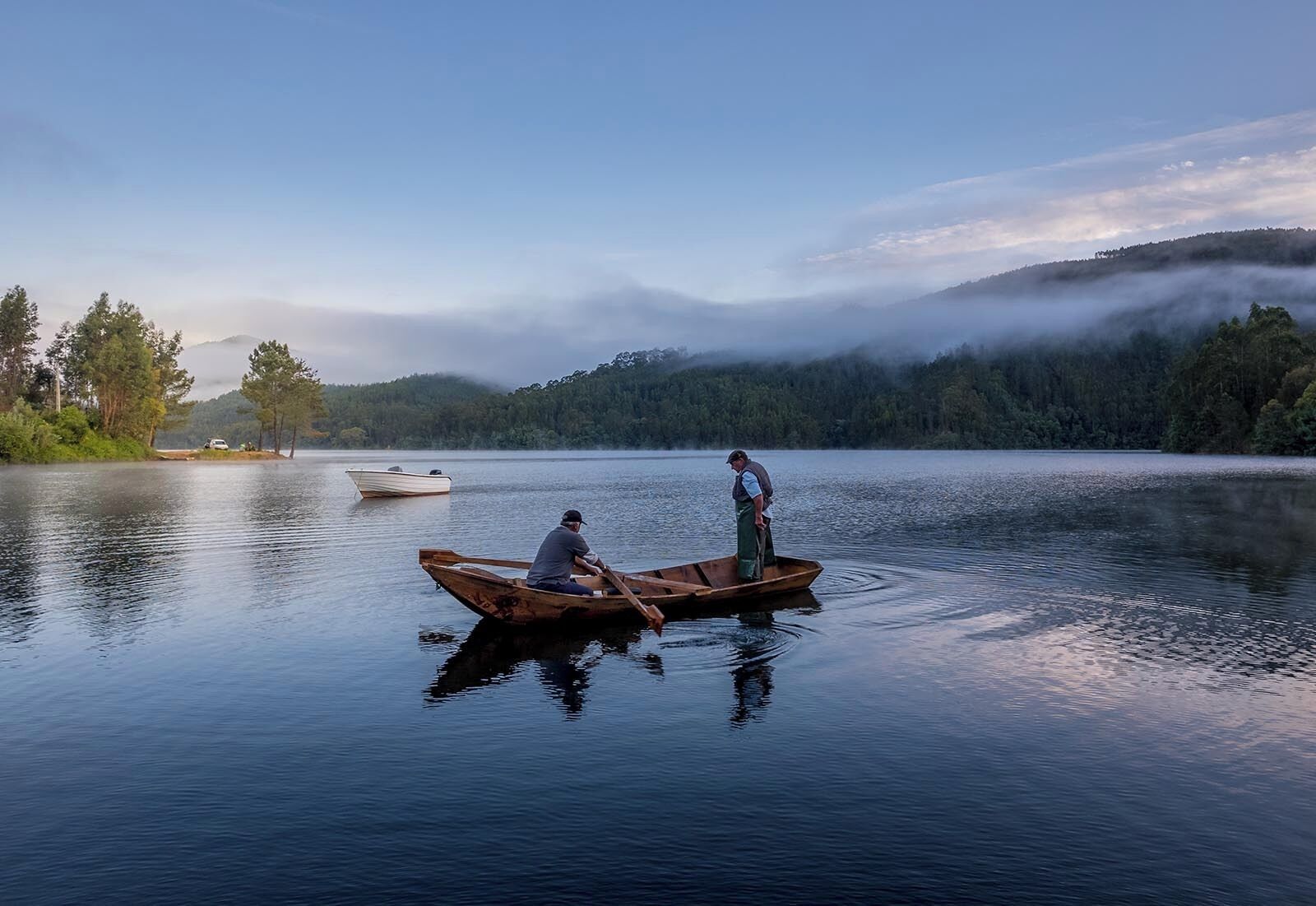 Un lac paisible où flottent deux bateaux en bois, l’un avec une personne qui rame et l’autre avec une personne debout.