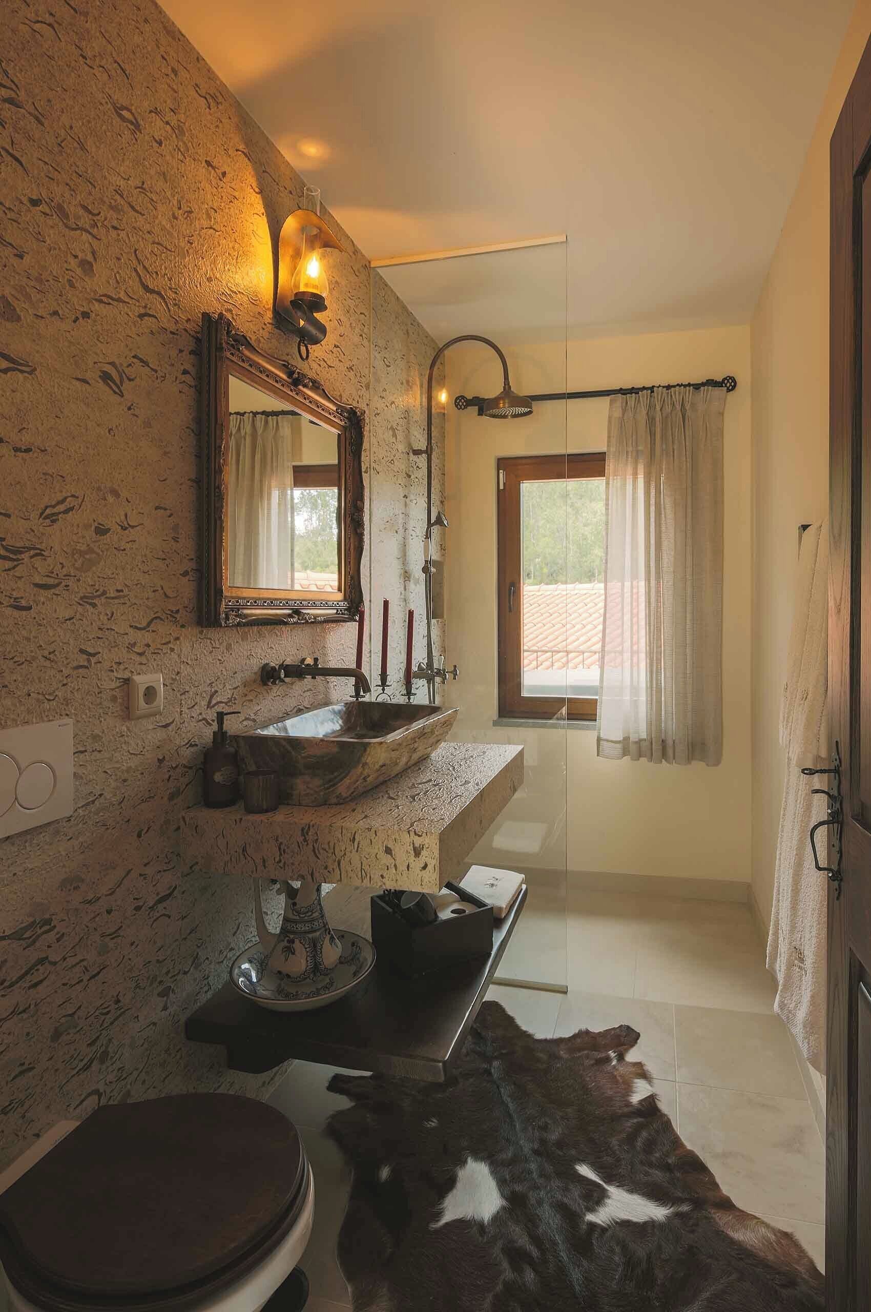 A bathroom with a rustic stone wall, a wooden vanity, a large mirror, a sink with a vessel basin, a shower with a handheld showerhead, and a window with curtains.