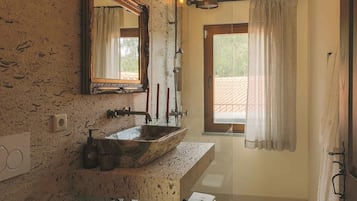A bathroom with a rustic stone wall, a wooden vanity, a large mirror, a sink with a vessel basin, a shower with a handheld showerhead, and a window with curtains.