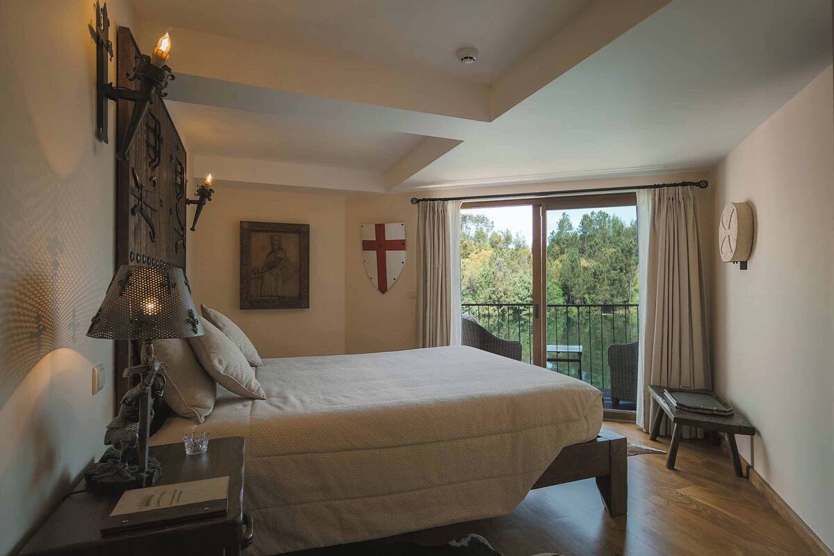 Une chambre à coucher avec un lit, une table de chevet, une lampe, une croix accrochée au mur et un balcon donnant sur des arbres.