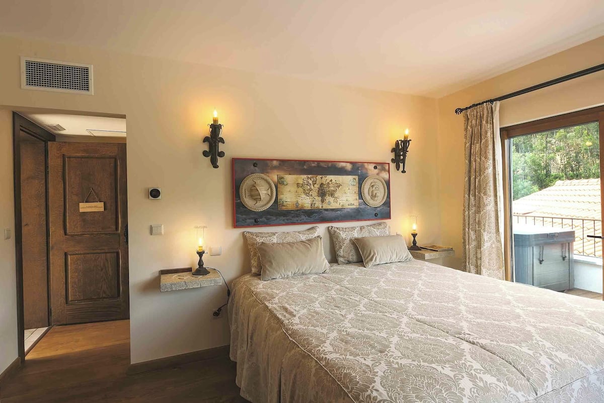 Une chambre avec un grand lit, deux appliques murales, un élément décoratif mural, une petite table avec une lampe et une vue sur la verdure par la fenêtre.