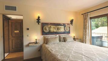Ein Schlafzimmer mit einem großen Bett, zwei Wandleuchten, einem Wanddekor, einem kleinen Tisch mit einer Lampe und Blick auf Grünpflanzen durch ein Fenster.