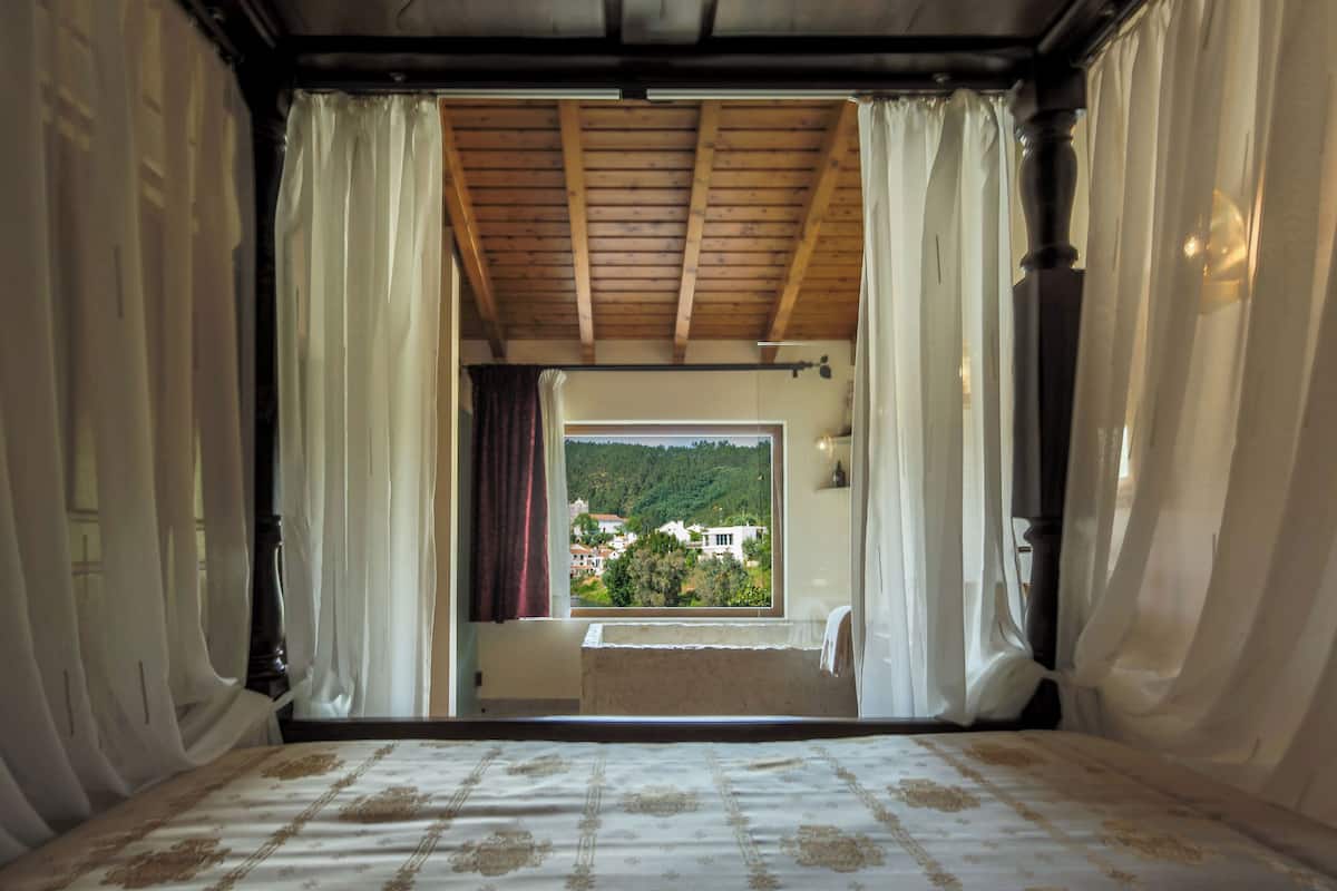 Une chambre avec un grand lit, une fenêtre avec des rideaux et une vue sur une maison et de la verdure à l’extérieur.