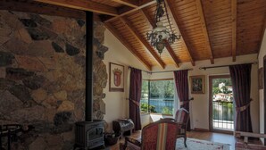 Une pièce chaleureuse aux murs de pierre, au plafond en bois, dotée d’une cheminée et de grandes fenêtres offrant une vue sur un lac.