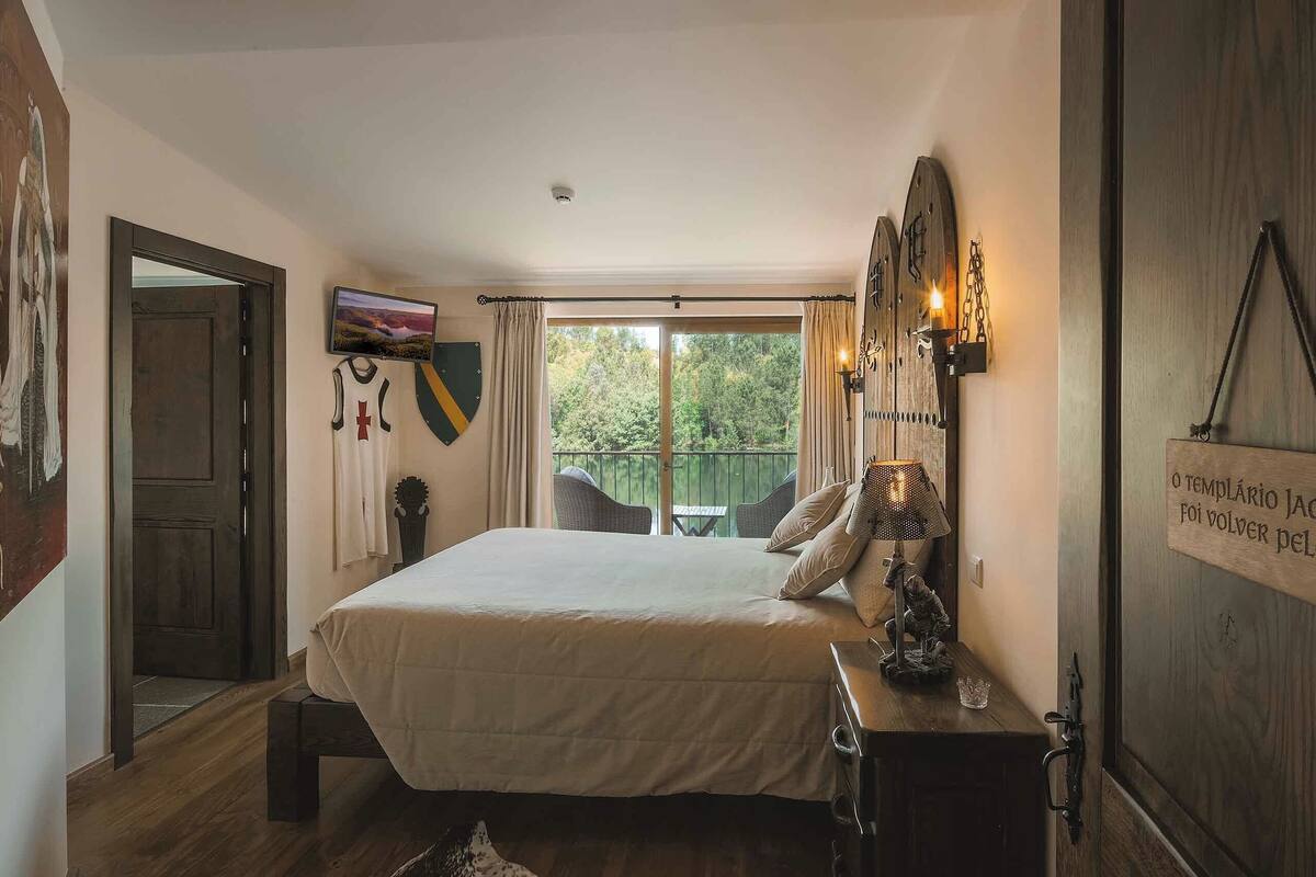 Une chambre avec un grand lit, une petite table avec une lampe, une porte en bois et une vue sur la verdure.
