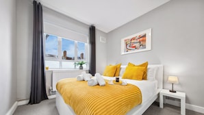 Una habitación de hotel moderna con una cama grande, una mesita de noche con lámpara, una ventana con cortinas y un cuadro en la pared.