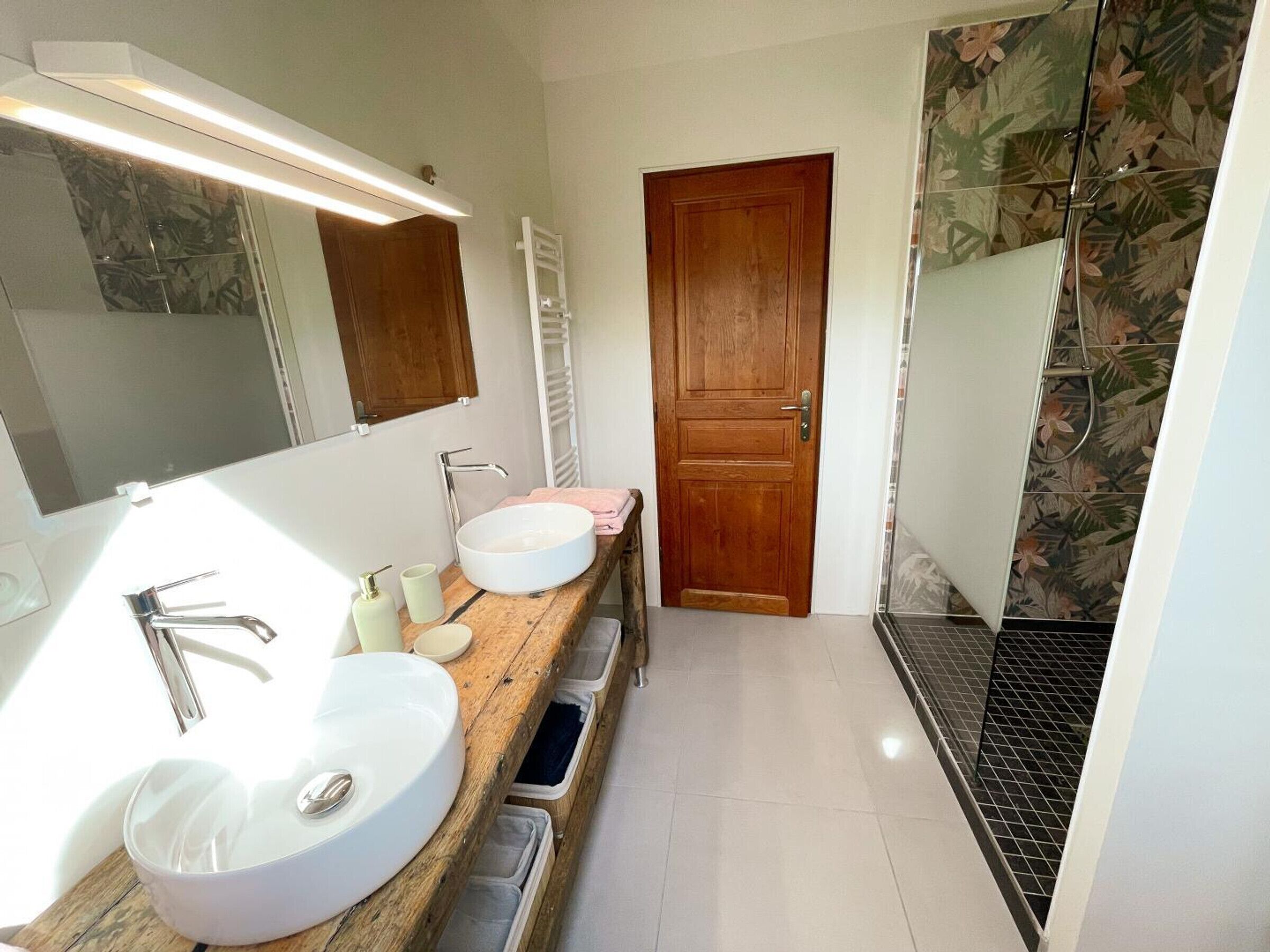 A bathroom with two sinks, a mirror, a wooden vanity, and a shower with a floral tile design.