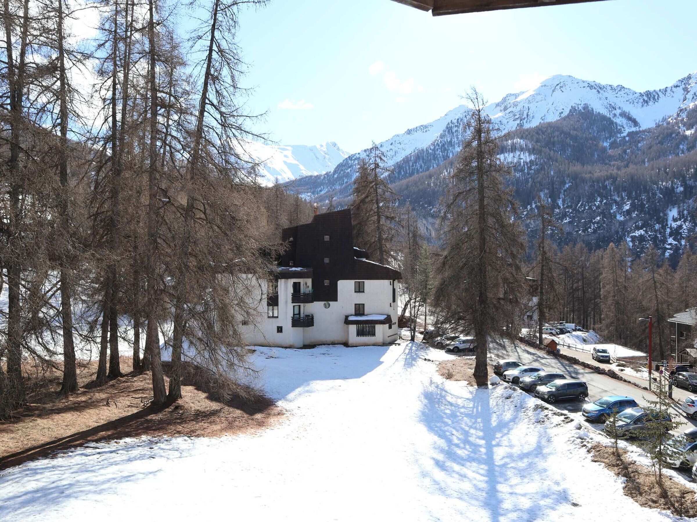 A white building with a black roof, surrounded by snow-covered ground and bare trees, with mountains in the background.