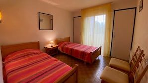 Une petite chambre agréable, dotée de deux lits simples, d’une table de chevet avec une lampe, d’une chaise et d’une fenêtre avec des rideaux.