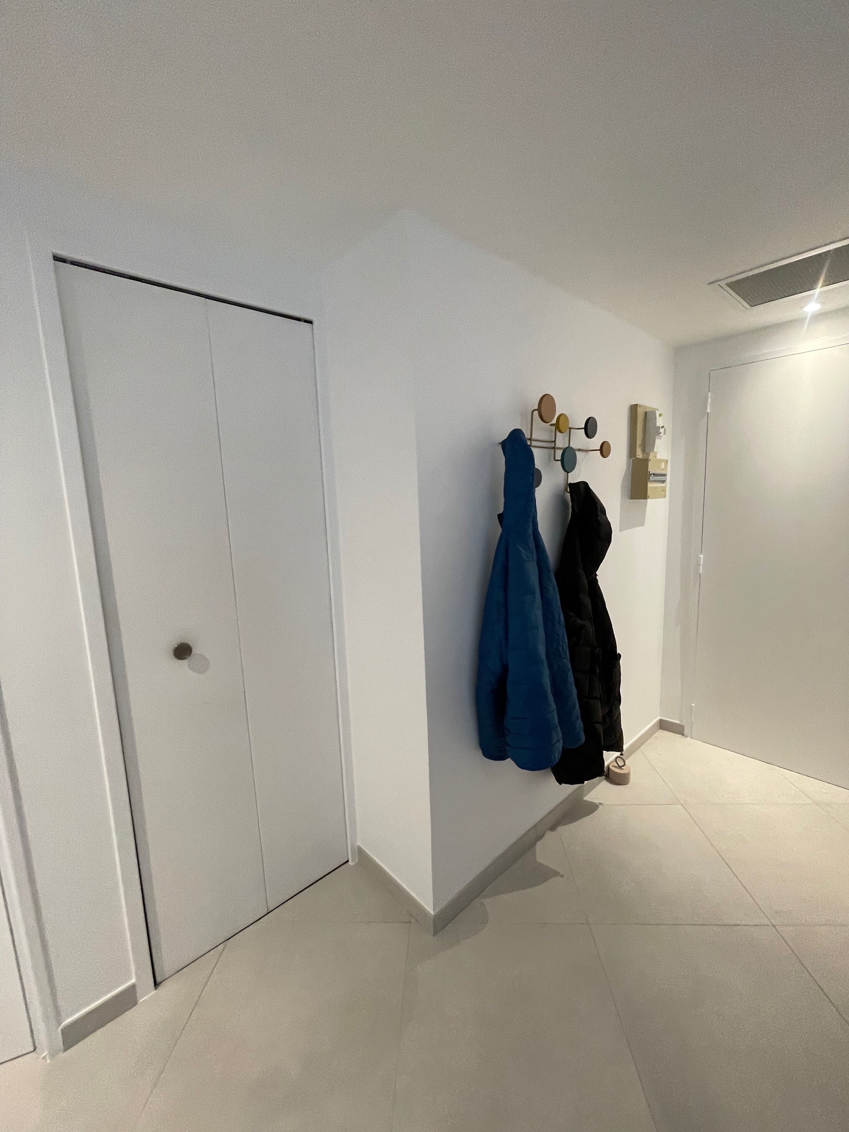 A hallway with white walls and a tiled floor. There are two doors, one closed and one open. A coat rack with several coats is mounted on the wall.
